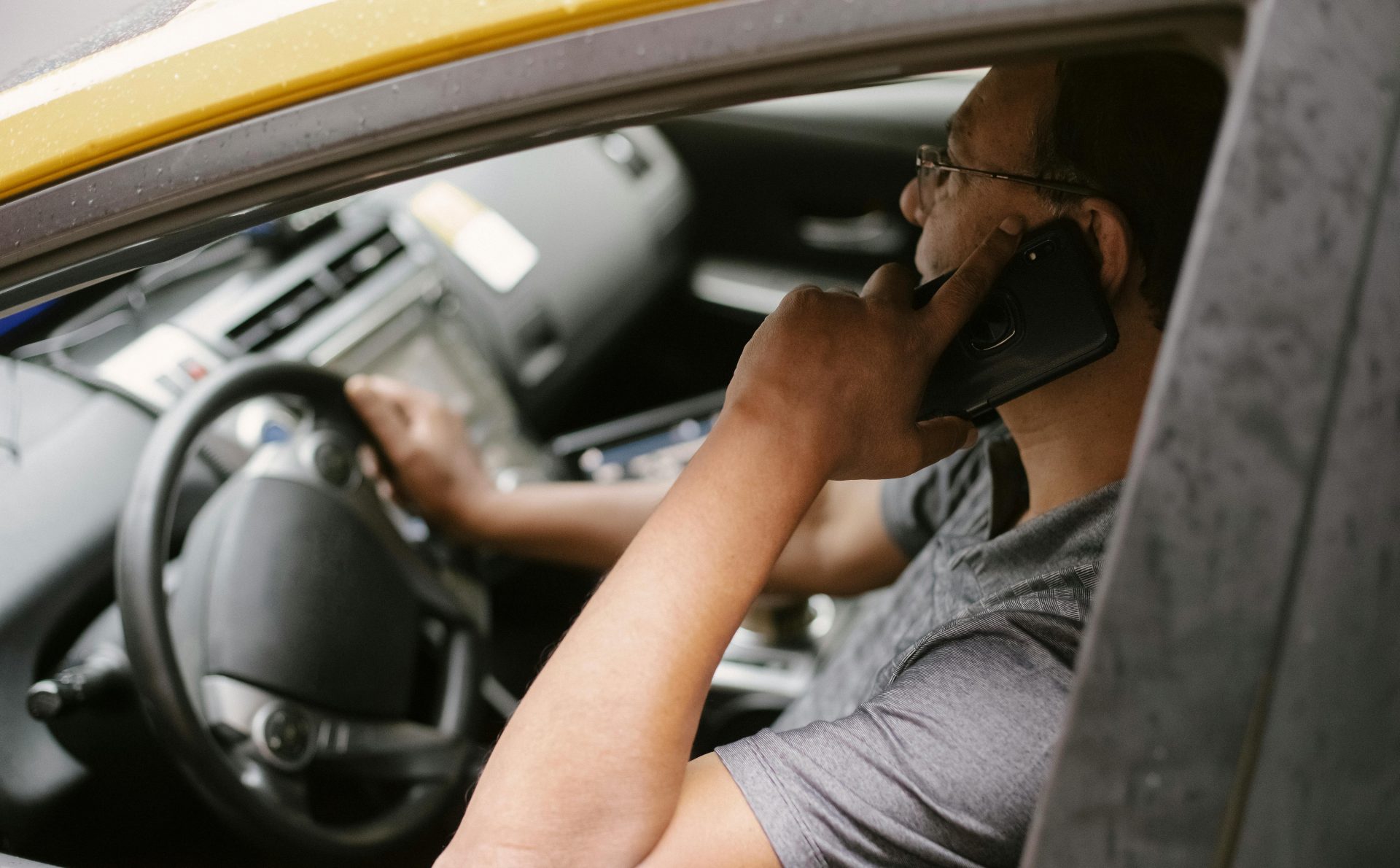 A male taxi driver in a yellow cab talks on the phone while driving in the city.